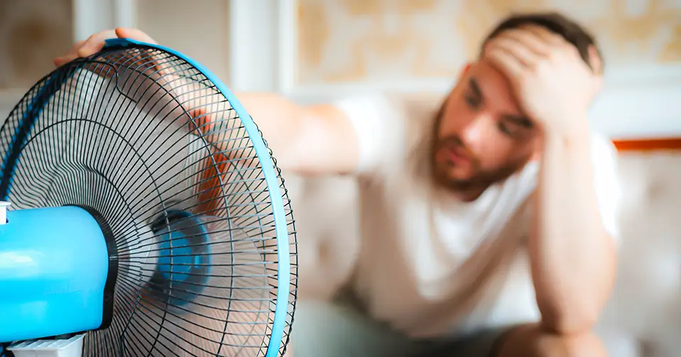 Man sits in front of a fan, because he's sweating that his AC is broken and needs repair.